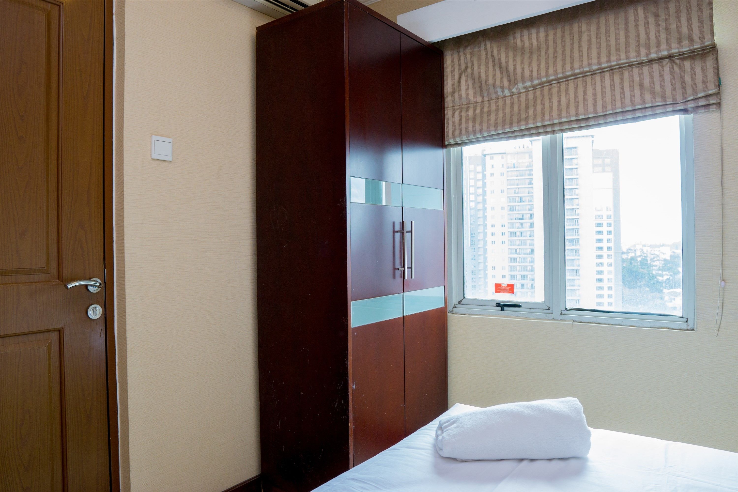 A hotel room with a wooden wardrobe, a bed with a white pillow, and a window with a view of high-rise buildings.