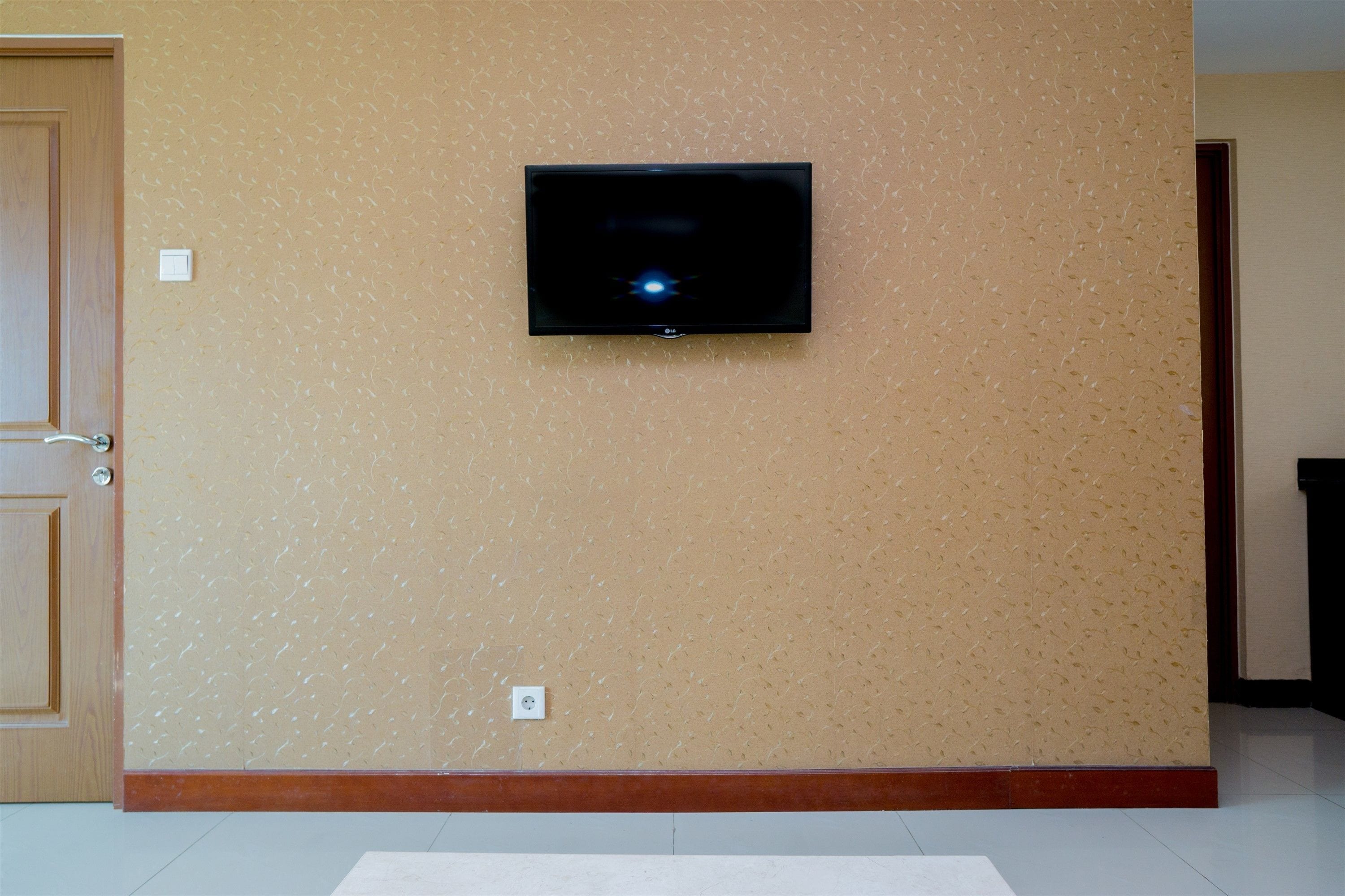 A room with a mounted flat-screen TV on the wall, a wooden door, and a light-colored floor.