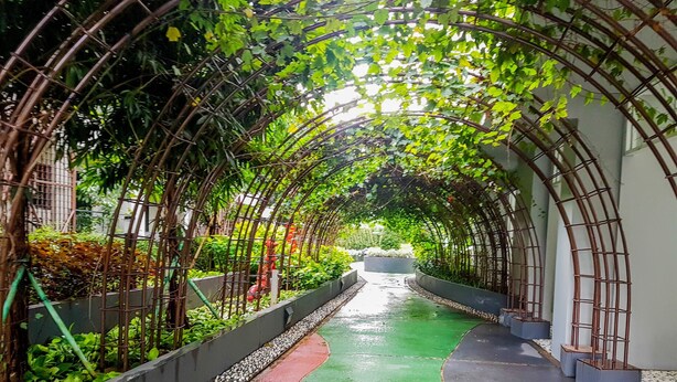 A covered pathway with a green and red surface, surrounded by lush greenery and a metal arch structure. - Nice & Comfy Studio Apartment Casa De Parco (Pagedangan)
