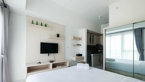 A modern hotel room with a large bed, a flat-screen TV, built-in shelves, a small kitchen area, and a glass door leading to a balcony. - Nice & Comfy Studio Apartment Casa De Parco (Pagedangan)