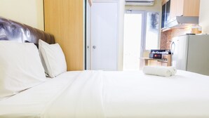 Ett hotellrum med en säng, en träpanel som huvudgavel, en vit dörr, ett litet pentry med ett kylskåp och ett fönster.
