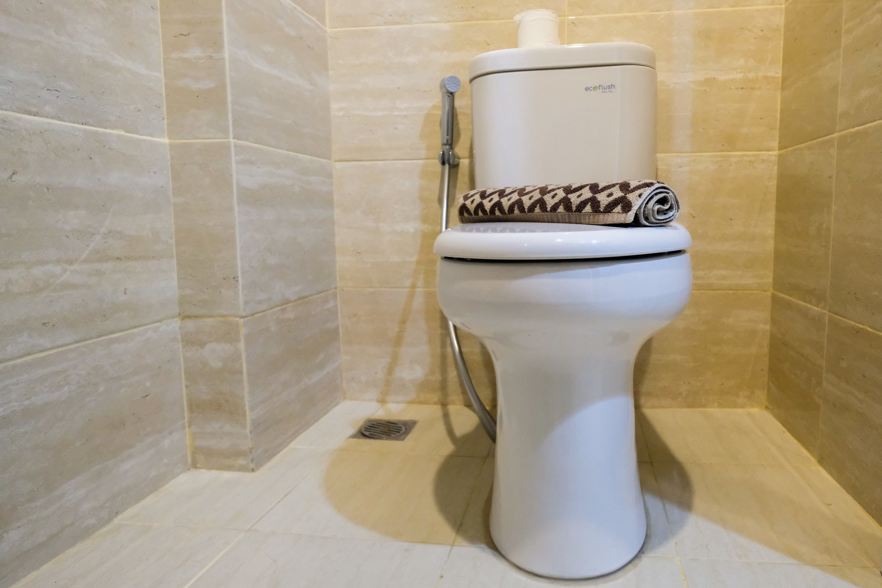 A toilet with a toilet paper holder and a bidet attachment in a bathroom with beige tiles.
