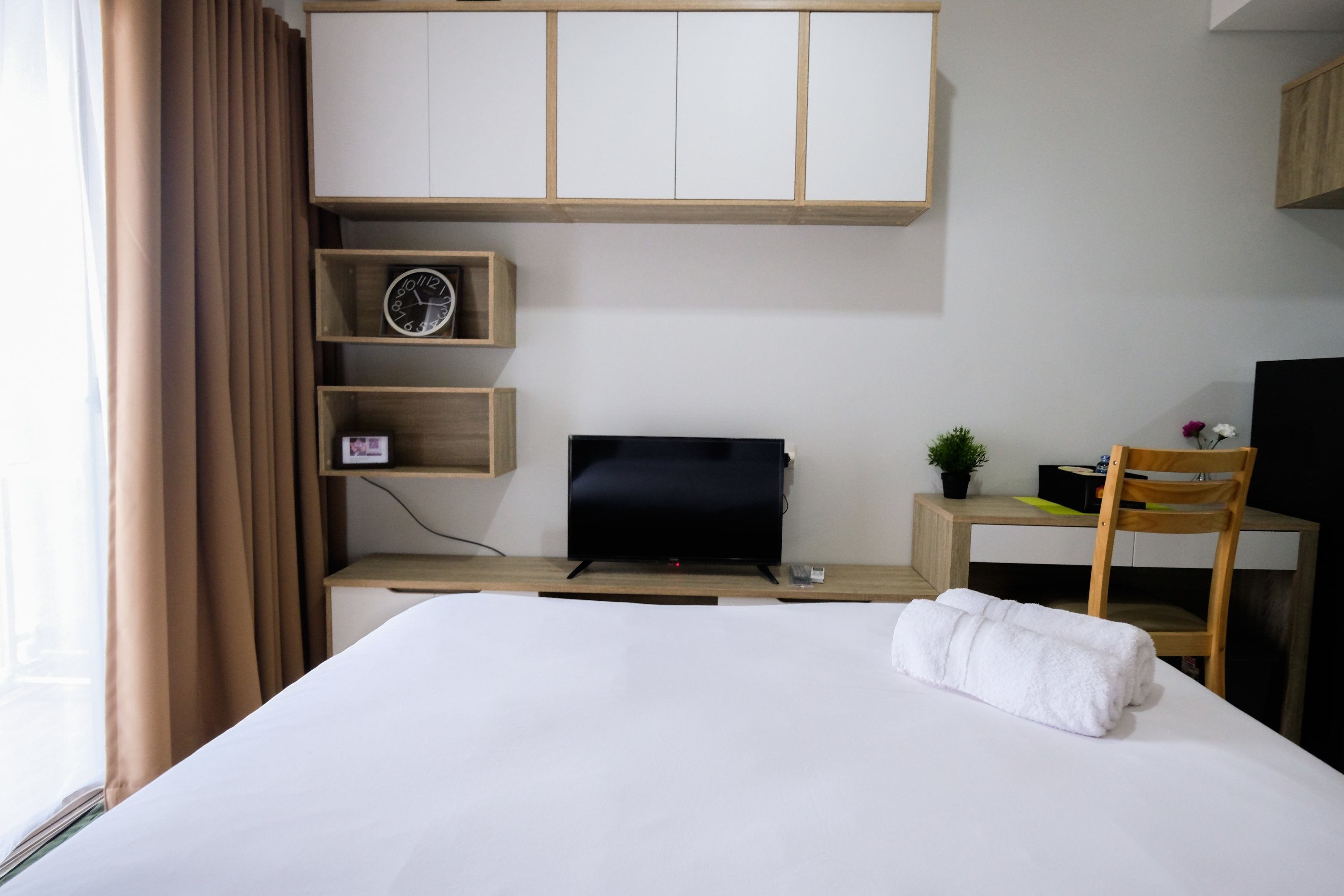 A hotel room with a bed, a desk with a computer, a TV, and a small shelf with a clock.