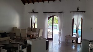 Un vaste espace de vie comprenant un canapé, des fauteuils, une table basse et une table à manger. Il y a trois fenêtres avec des rideaux, une porte donnant sur l’extérieur et une vue sur une piscine.