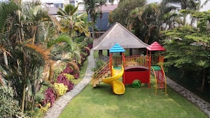 Un terrain de jeux équipé d’un toboggan et de structures d’escalade, entouré d’un jardin agrémenté de palmiers et de diverses plantes.