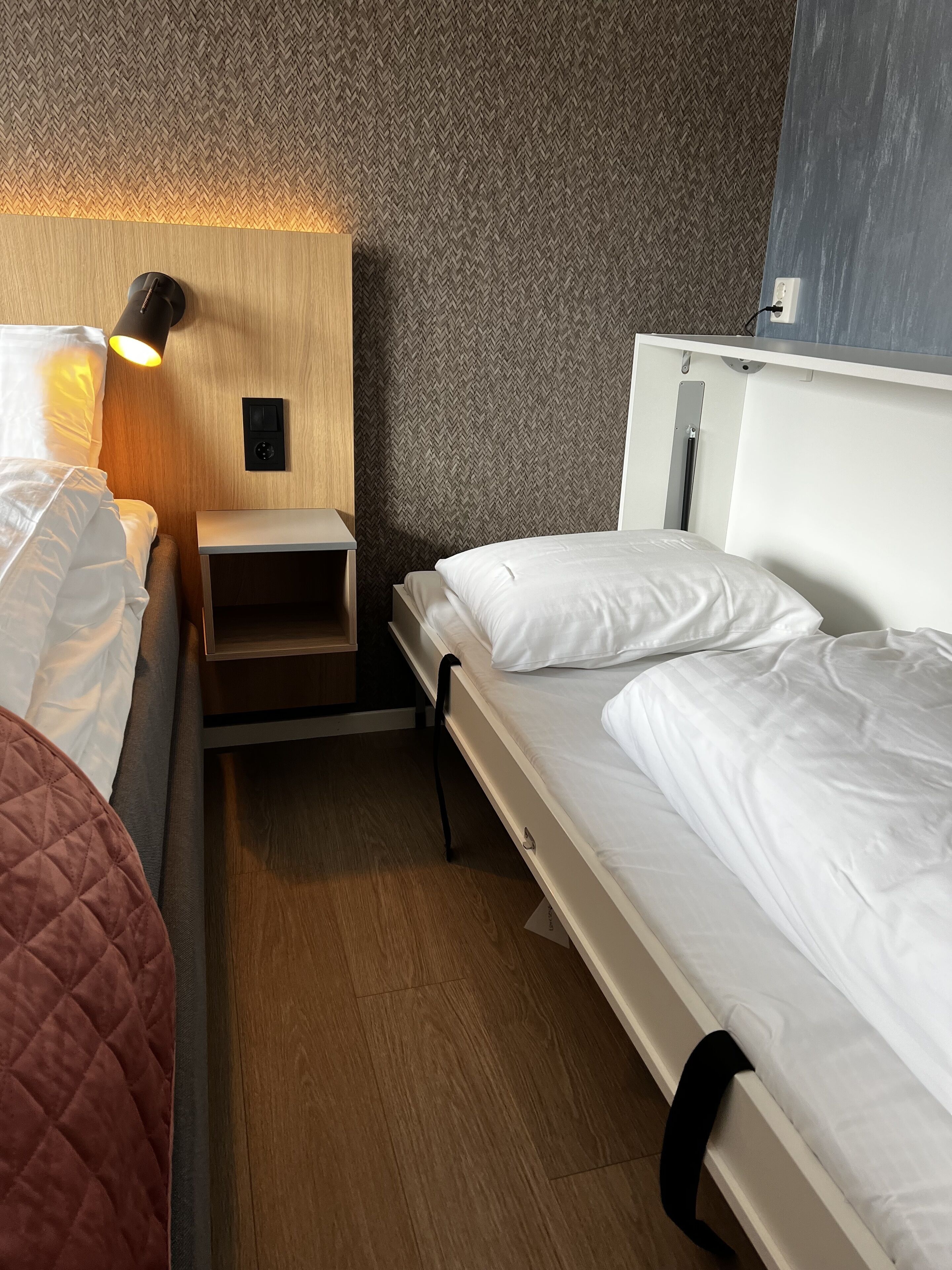 Ein kleines Hotelzimmer mit einem hölzernen Kopfteil, einem Nachttisch mit Lampe und einem Bett mit weißer Bettwäsche.