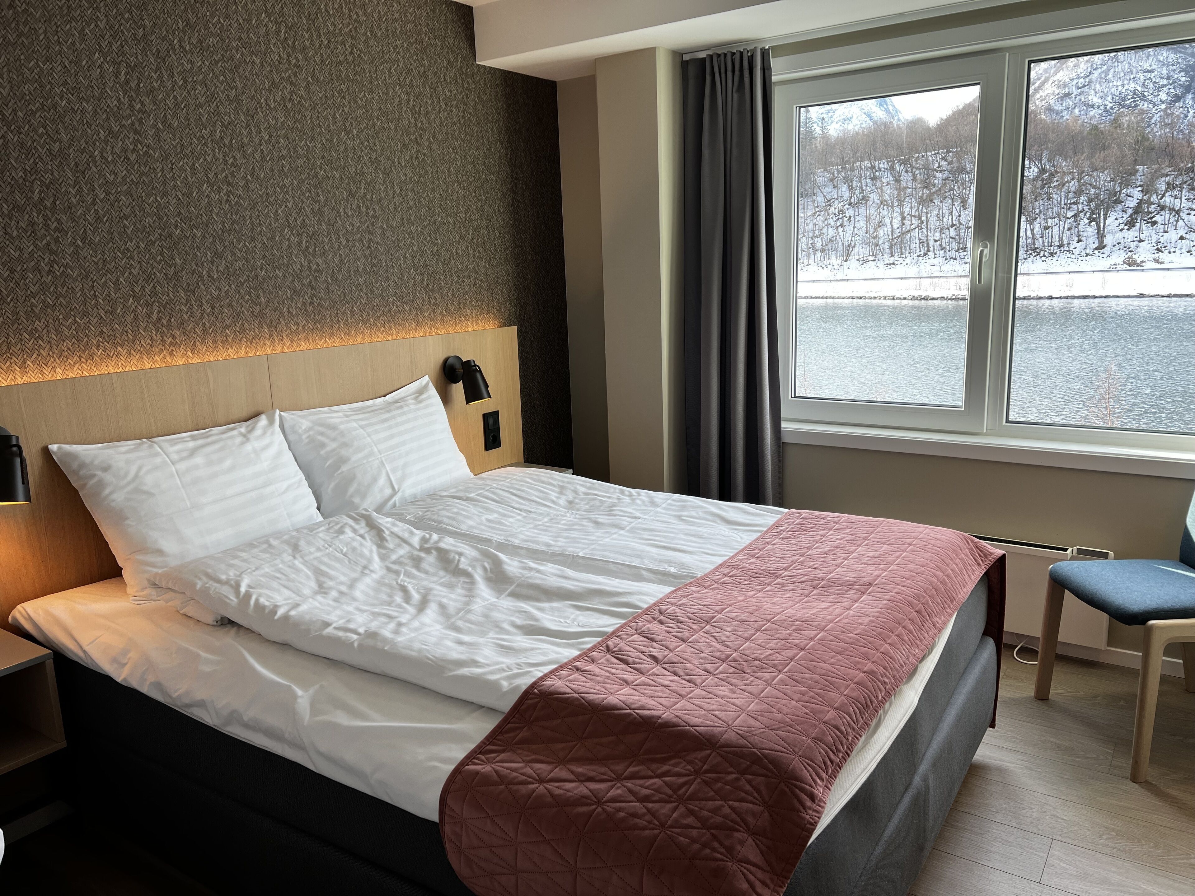 A neatly made bed with white linens, a pink quilted throw, and a headboard with a light fixture.