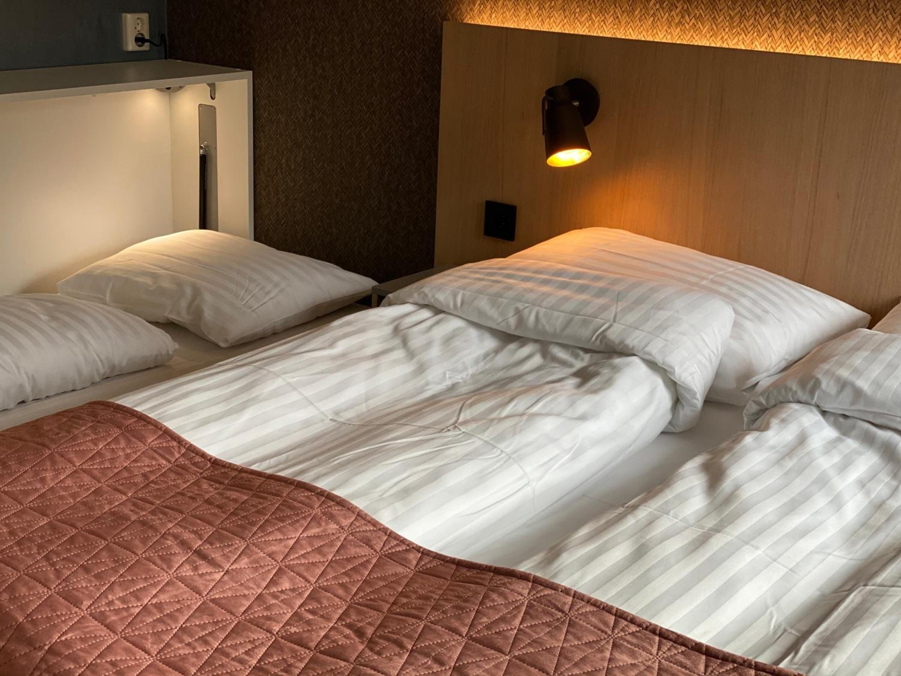 Un lit bien fait, avec du linge de lit blanc et une couverture matelassée marron, un luminaire fixé au mur et un mur à texture.