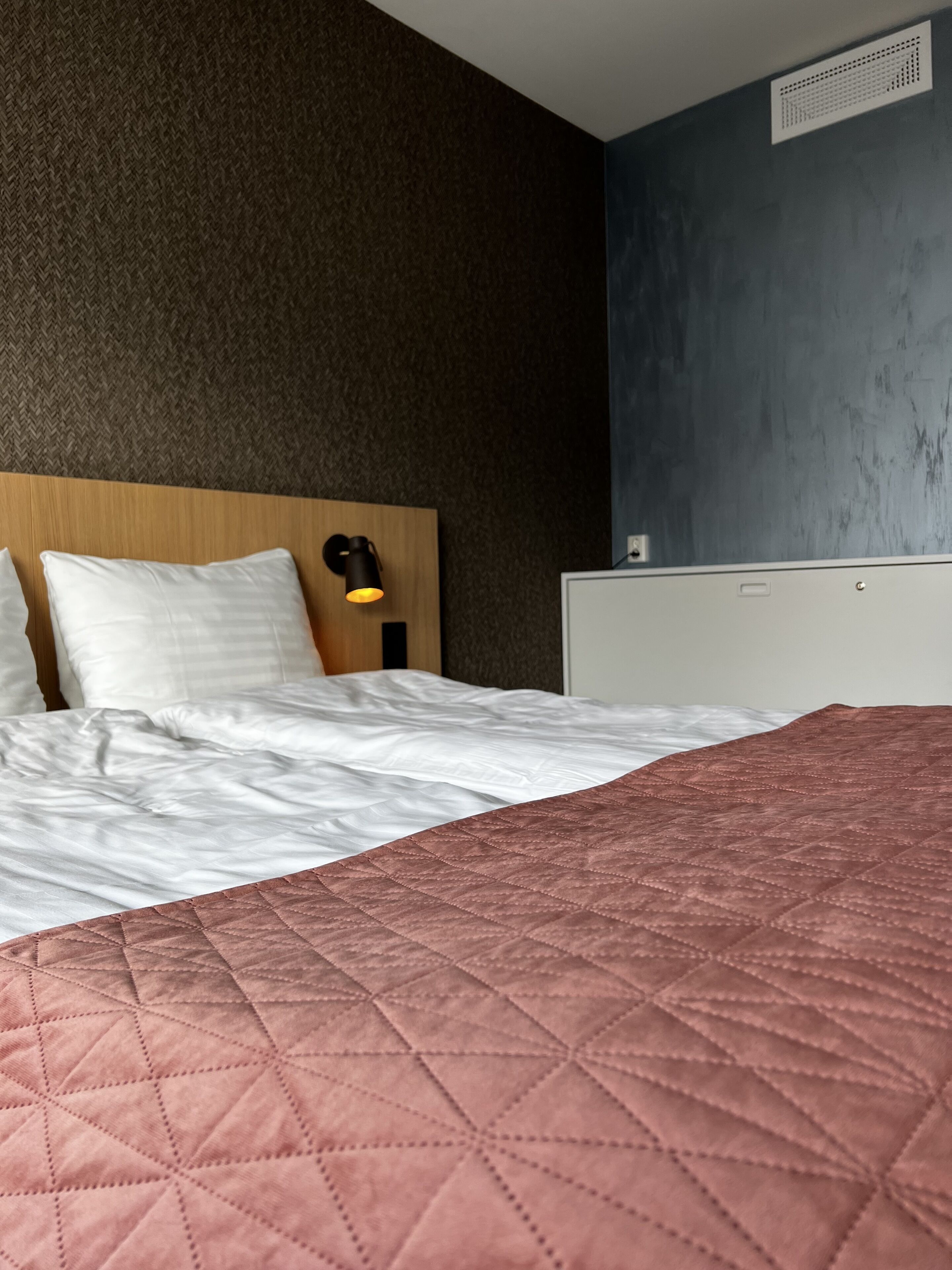 A hotel room with a bed, a nightstand, a wall-mounted lamp, and a ventilation unit.
