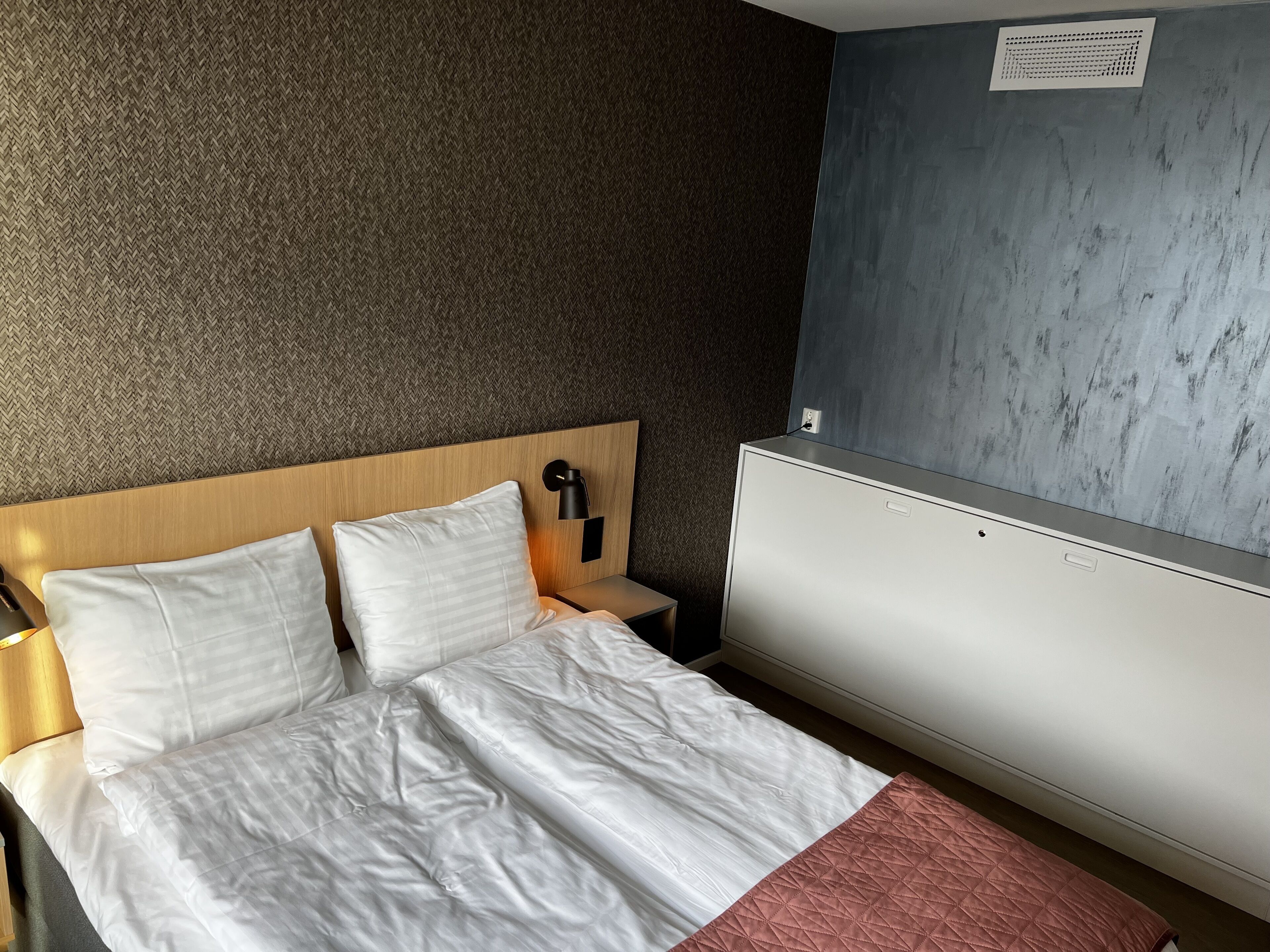 Ein Hotelzimmer mit Bett, Nachttisch, Lampe und einer Wand-Klimaanlage.