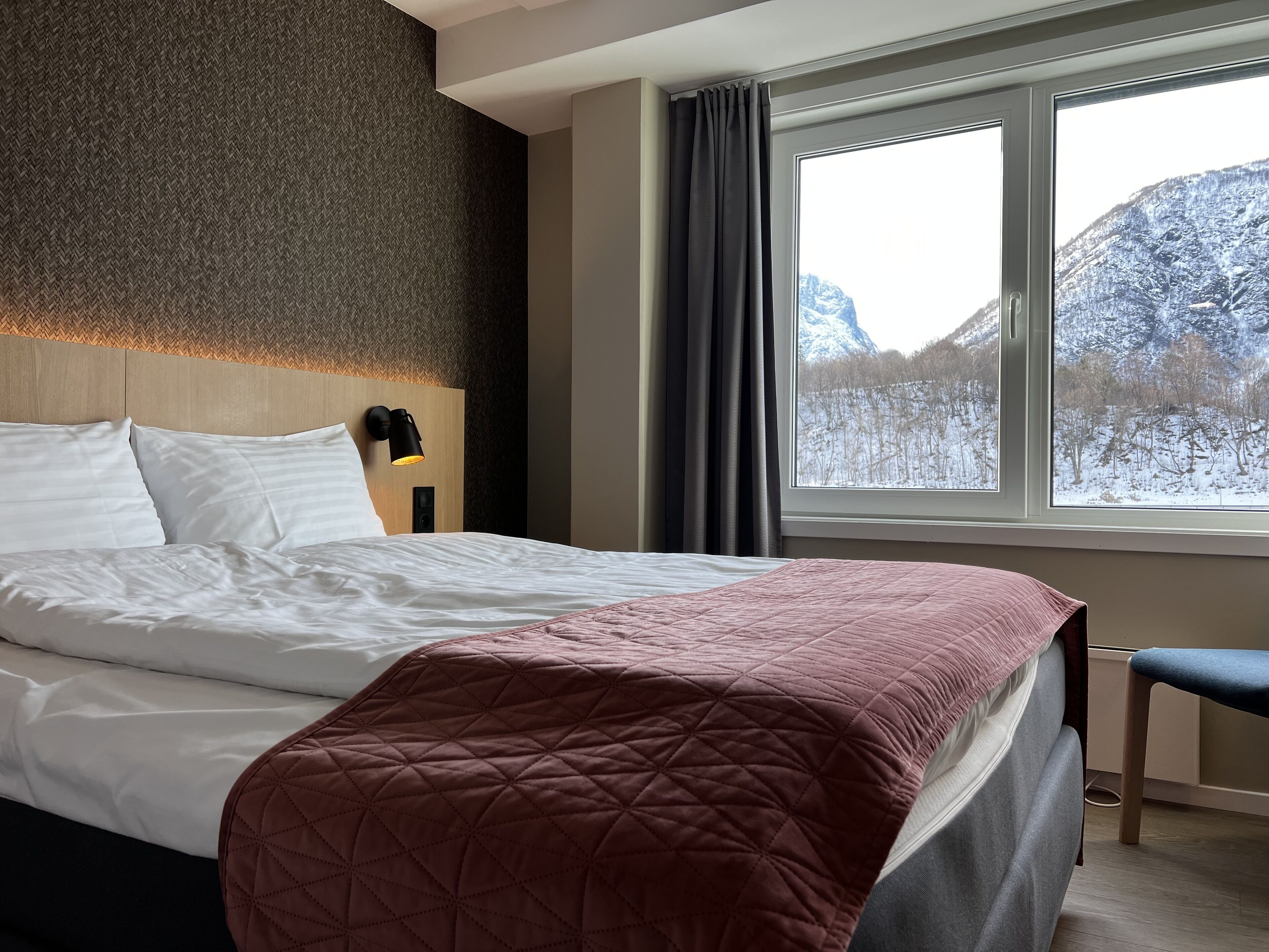 A hotel room with a bed, a headboard, a nightstand with a lamp, a window with a view of snow-covered mountains, and a bench.