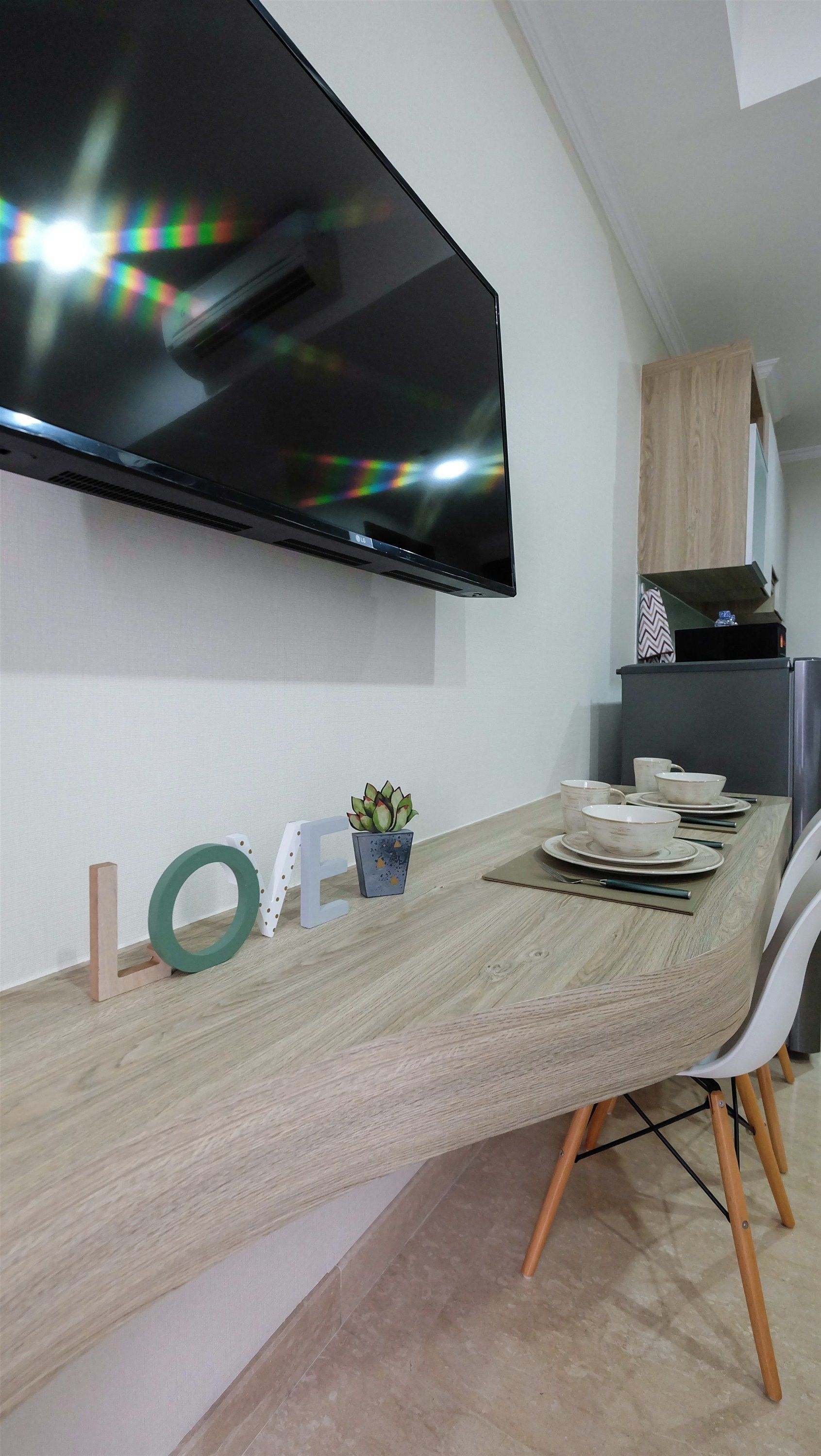A modern kitchen with a flat-screen TV mounted above a wooden dining table, a small potted plant, and a refrigerator.