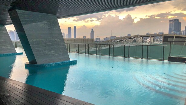 Une piscine intérieure offrant une vue sur les toits de la ville, une terrasse en bois et une structure en béton au-dessus.