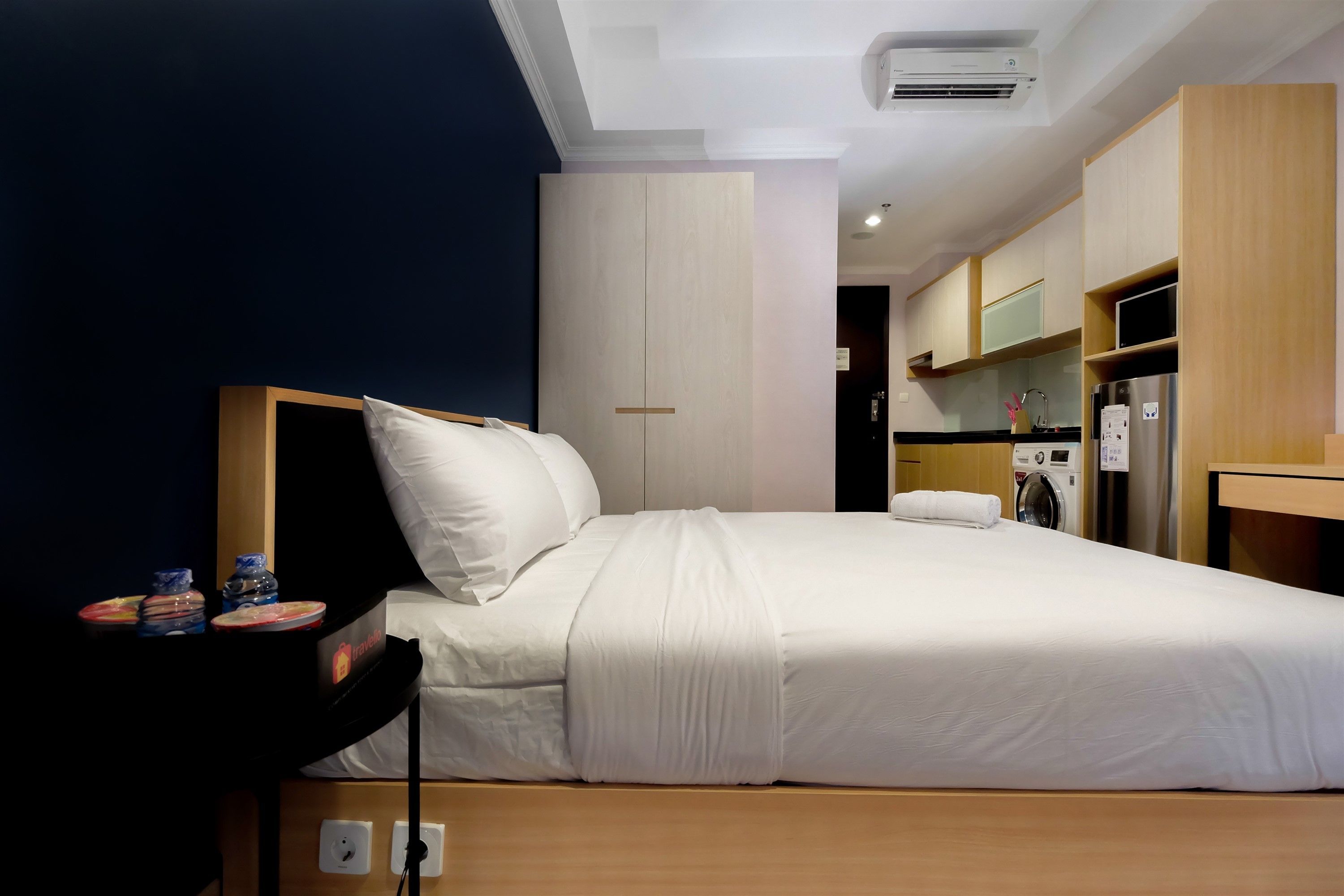 Quarto de hotel com cama, uma pequena mesa com objetos, máquina de lavar e cozinha compacta.