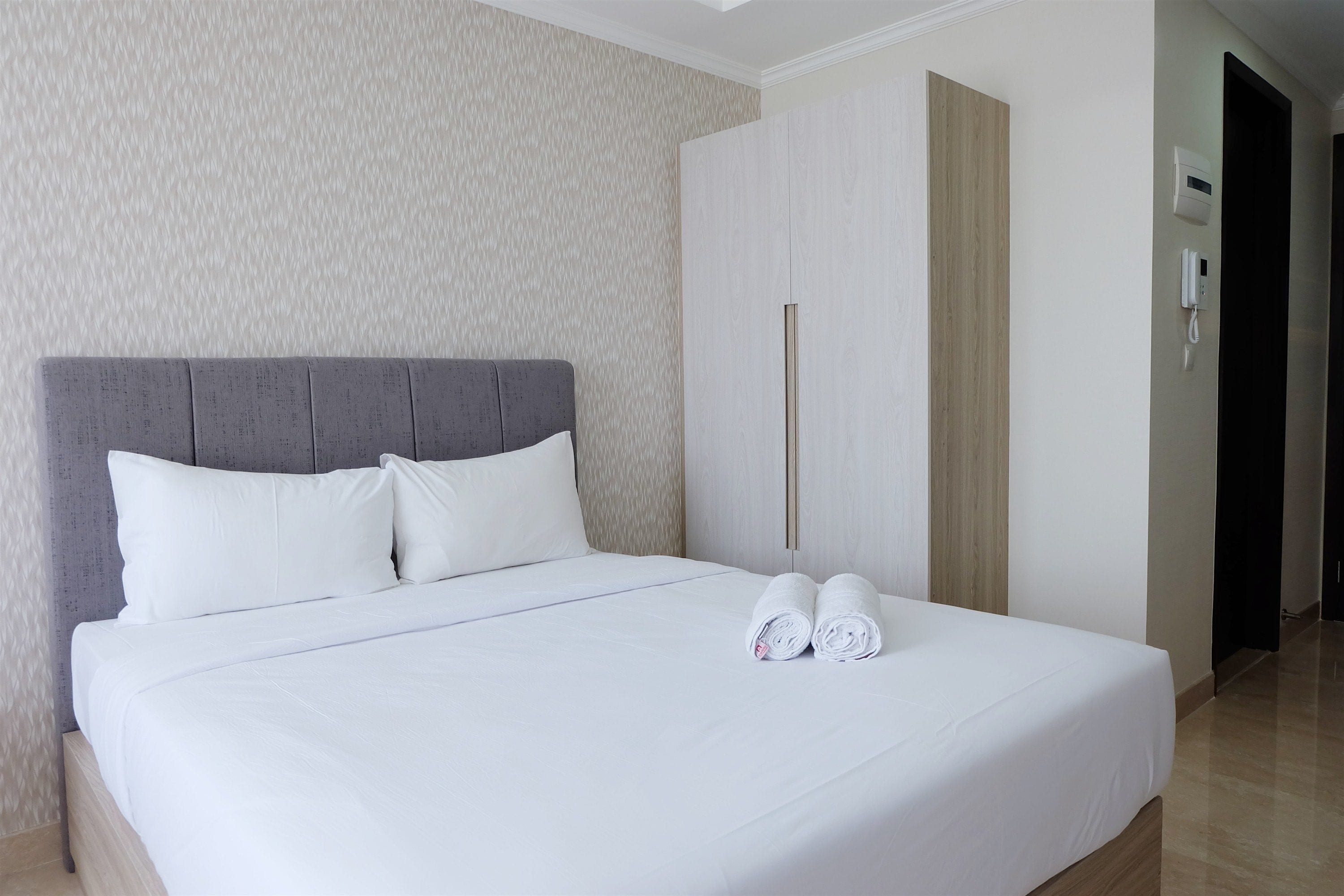 A neatly made bed with white linens and pillows, a grey headboard, and a pair of rolled towels on the bed.