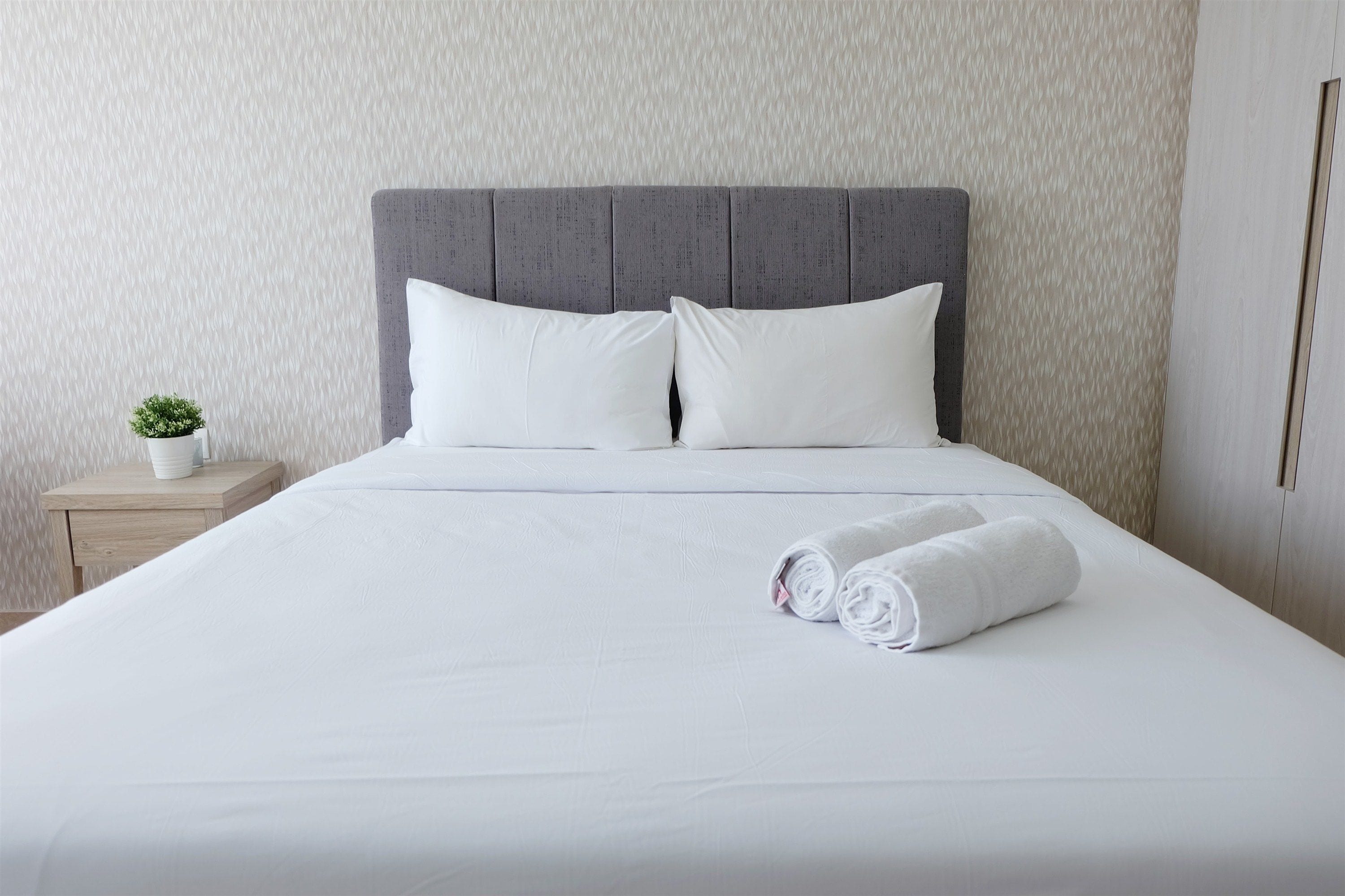 A neatly made bed with white linens and pillows, a small bedside plant, and a wooden nightstand.