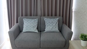 A grey sofa with patterned cushions, a wooden side table with a potted plant, and a curtain backdrop. - Comfy Studio Menteng Park Apartment (Jakarta)