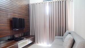 A modern living room with a wooden panel wall, a flat-screen TV, a grey sofa, and a window with curtains. - Great Location 2BR Maple Park Apartment (Jakarta)
