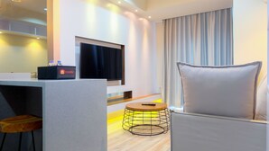 Moderni hotellihuone, jossa on seinälle asennettu taulutelevisio, harmaa sohva, pieni pyöreä pöytä, jonka jalassa on metallirunko, sekä minibaari, jossa on Travelion logo.