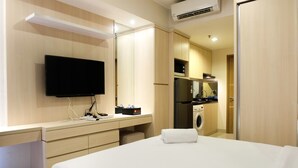 A modern hotel room with a flat-screen TV, a desk with a computer, a washing machine, and a bed with a white towel. - Artistic Studio Apartment at The Oasis Cikarang (Cikarang)