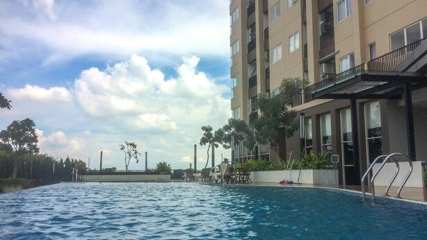 A swimming pool with a view of a multi-story building, surrounded by greenery and a partly cloudy sky. - Artistic Studio Apartment at The Oasis Cikarang (Cikarang)