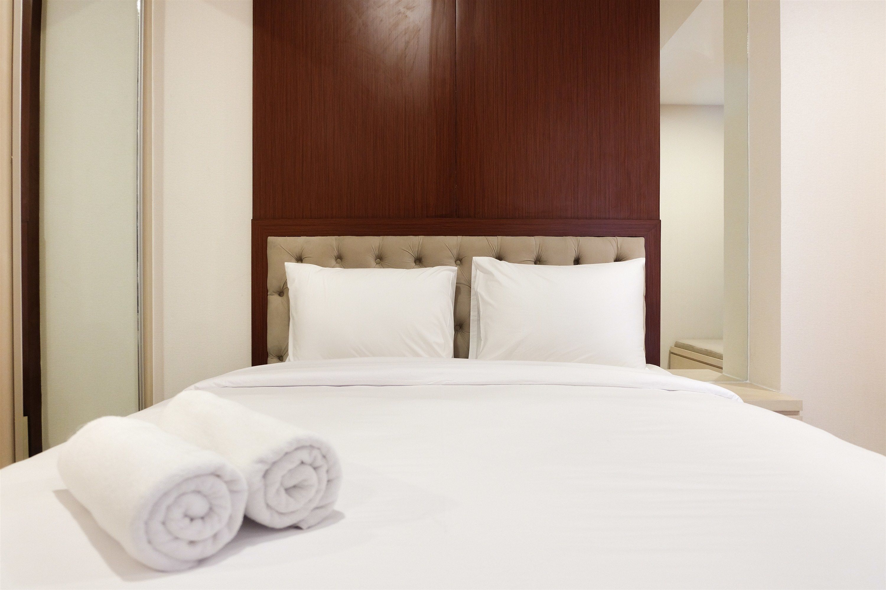 A neatly made bed with white linens and two rolled towels, a wooden headboard, and a plain wall.