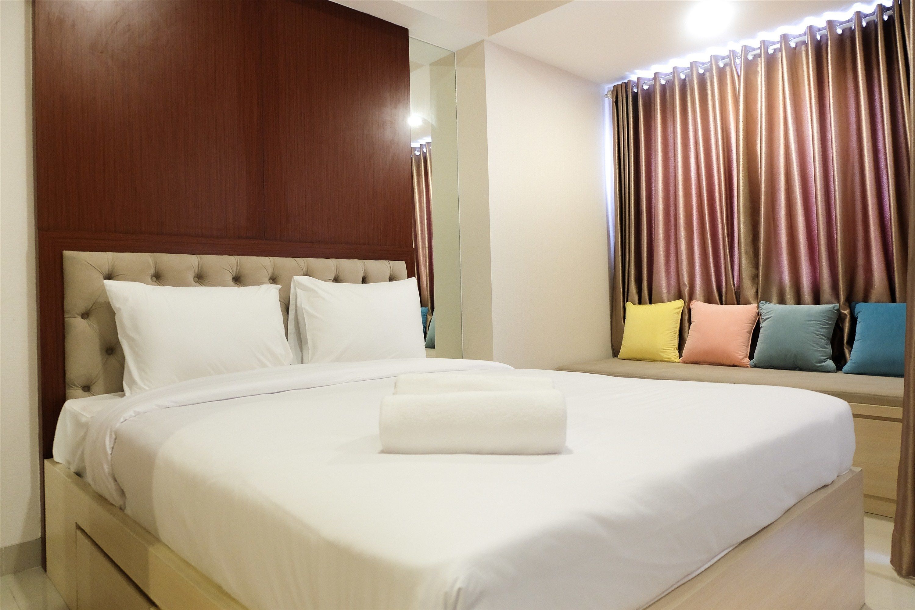 A neatly made bed with a white duvet and a rolled white towel in the center, a wooden headboard, and a seating area with colorful cushions.
