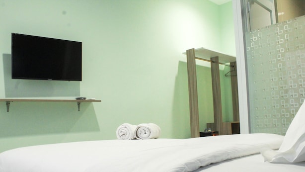 A hotel room with a bed, a flat-screen TV mounted on the wall, a towel rack with towels, and a shower area with frosted glass doors. - Studio Guesthouse Way Seputih Luxury near Central Park (Jakarta)