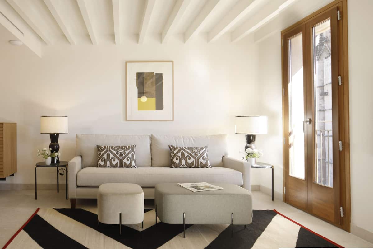 Un salon moderne avec un canapé, un pouf et des tables d’appoint avec des lampes. On y trouve également un meuble en bois et un tableau encadré accroché au mur.