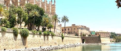 A historic building with a stone facade and multiple towers, adjacent to a calm river with a stone wall and palm trees.