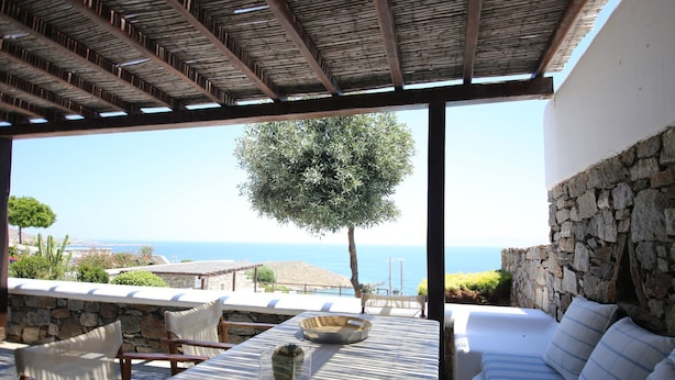A patio with a table and chairs, a stone wall, and a view of the sea. - Villa Attalus 3 (Mykonos)