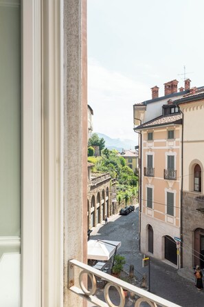 Une vue depuis un balcon donnant sur une rue pavée, bordée de bâtiments historiques, avec un ciel dégagé et des montagnes à l’horizon.