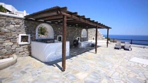 Un área de estar al aire libre con paredes de piedra, una pérgola, suelo de piedra y vista al mar.