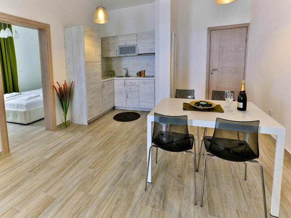 A modern kitchen with white cabinets, a microwave, and a sink. There is a dining table with four chairs and a bottle of wine.