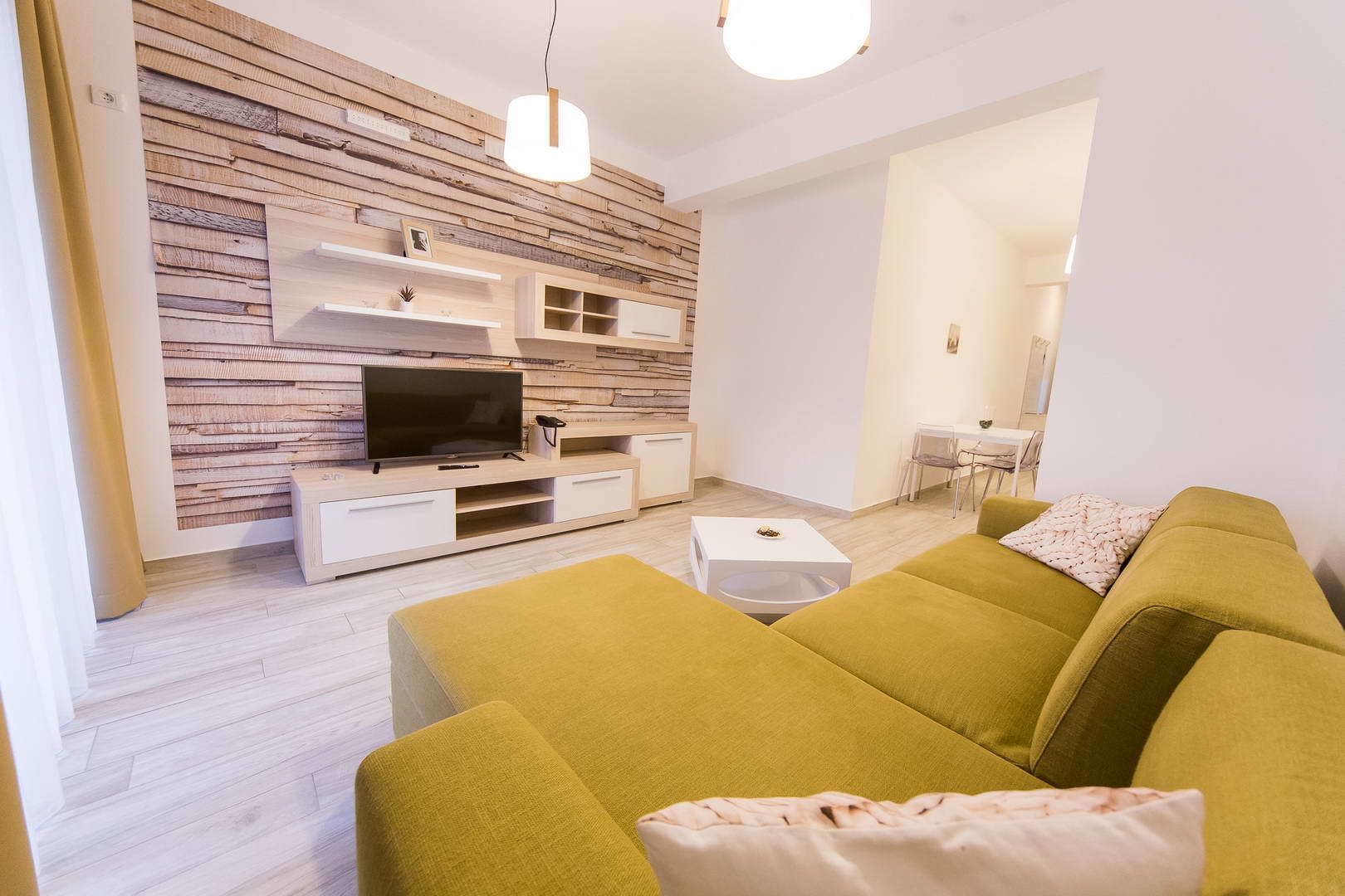Un salon moderne avec un mur recouvert de panneaux en bois, un téléviseur à écran plat, un canapé jaune et un coin repas à l’arrière-plan.
