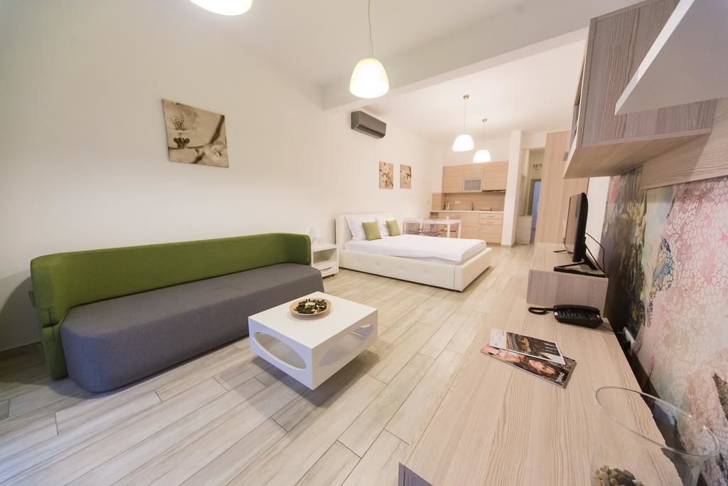 A modern living room with a green sofa, a white bed, and a small white coffee table. There is a kitchen area in the background with wooden cabinets and a microwave.