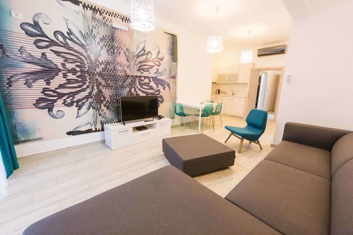 A modern living room with a large flat-screen TV, a sofa, a coffee table, and a dining area with chairs.