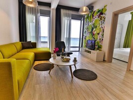 Uma sala de estar moderna com um sofá amarelo, uma mesa de centro redonda e uma televisão sobre um suporte.
