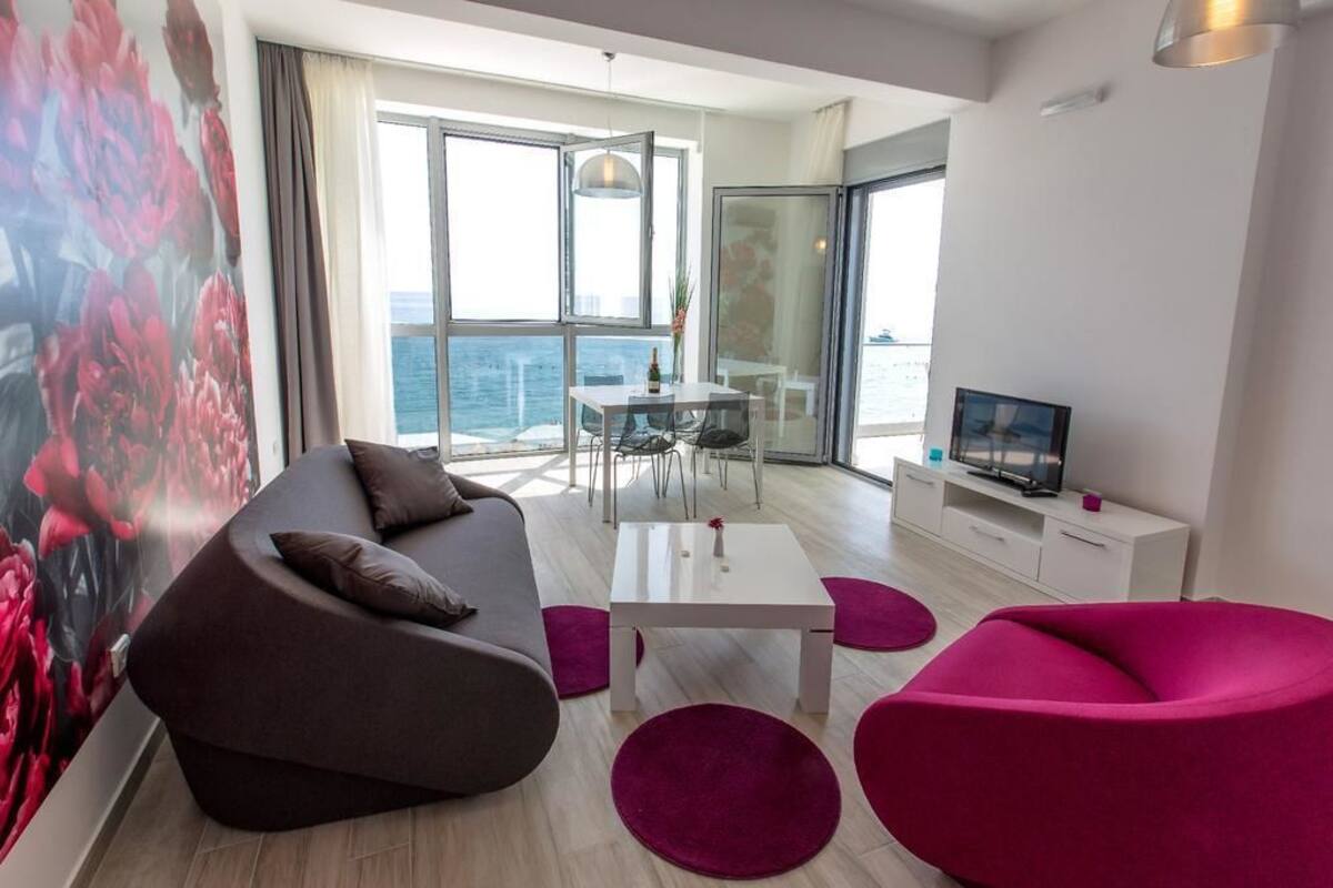 A modern living room with a sofa, a coffee table, and a TV. There is a balcony with a view of the sea.