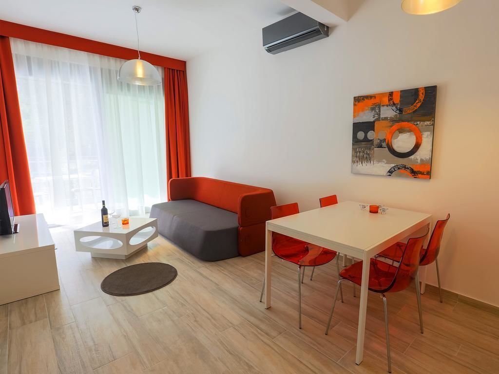 A modern living room with a sofa, dining table, and chairs. There is a painting on the wall and an air conditioning unit above the sofa.