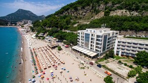 A beach with rows of umbrellas and sun loungers, a large building with multiple floors, and a mountainous backdrop. - Apart Hotel Sea Fort (Sutomore)