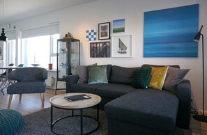 A modern living room with a grey sectional sofa, a round coffee table, and a bookshelf. There are framed pictures on the wall and a large abstract painting.