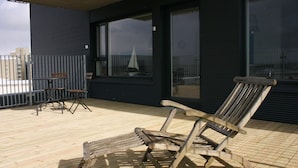 A wooden deck with a single wooden chair and a small table, a metal railing, and a building with large windows in the background. - Nest Apartment Sparrow (Reykjavik)