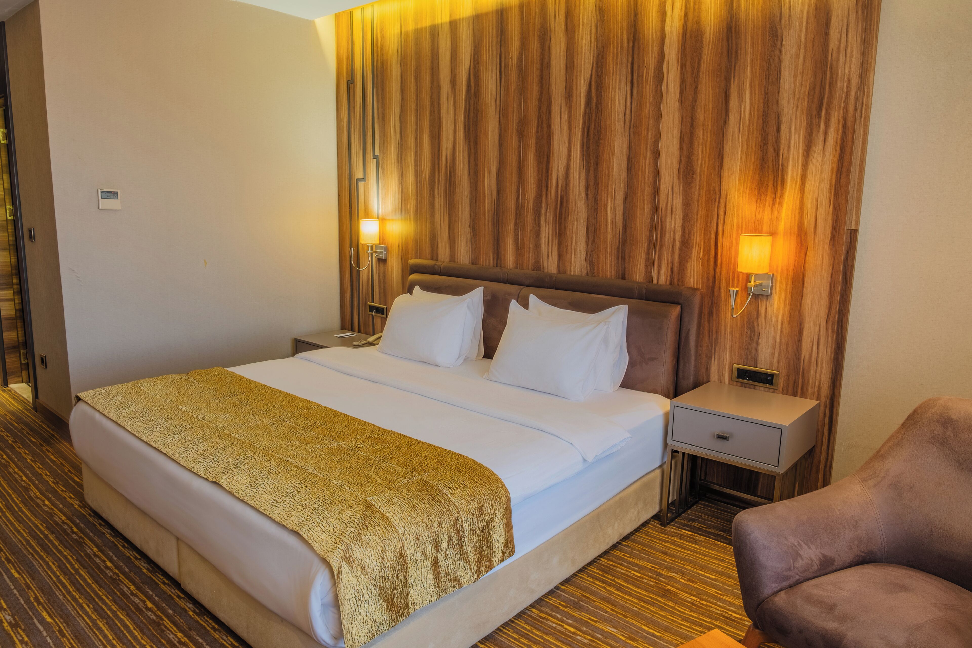 A hotel room with a large bed, two bedside tables, a nightstand, a chair, and a wooden wall paneling.
