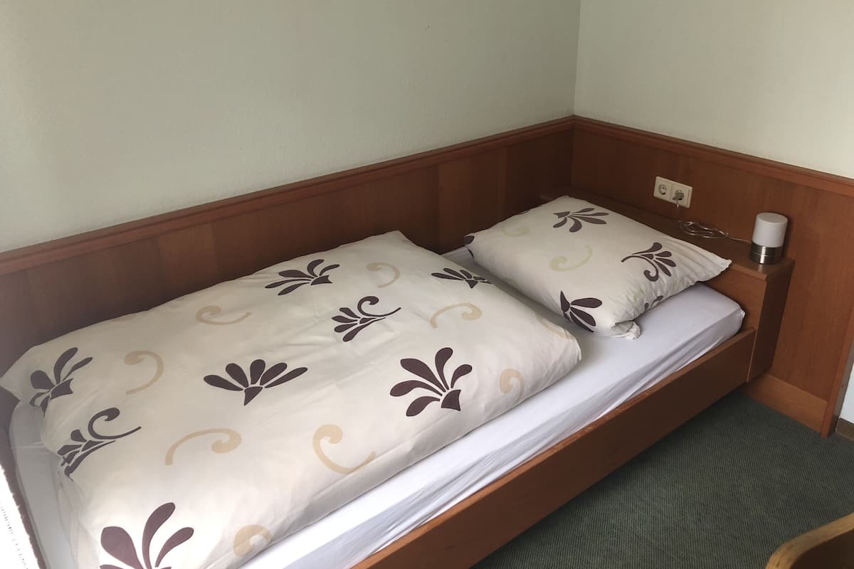 A single bed with a wooden headboard, two pillows with a floral pattern, and a bedside table with a lamp.