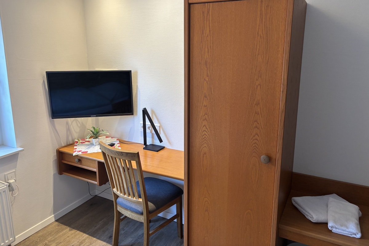 A compact room with a wooden desk, a chair, a television mounted on the wall, and a wooden wardrobe.