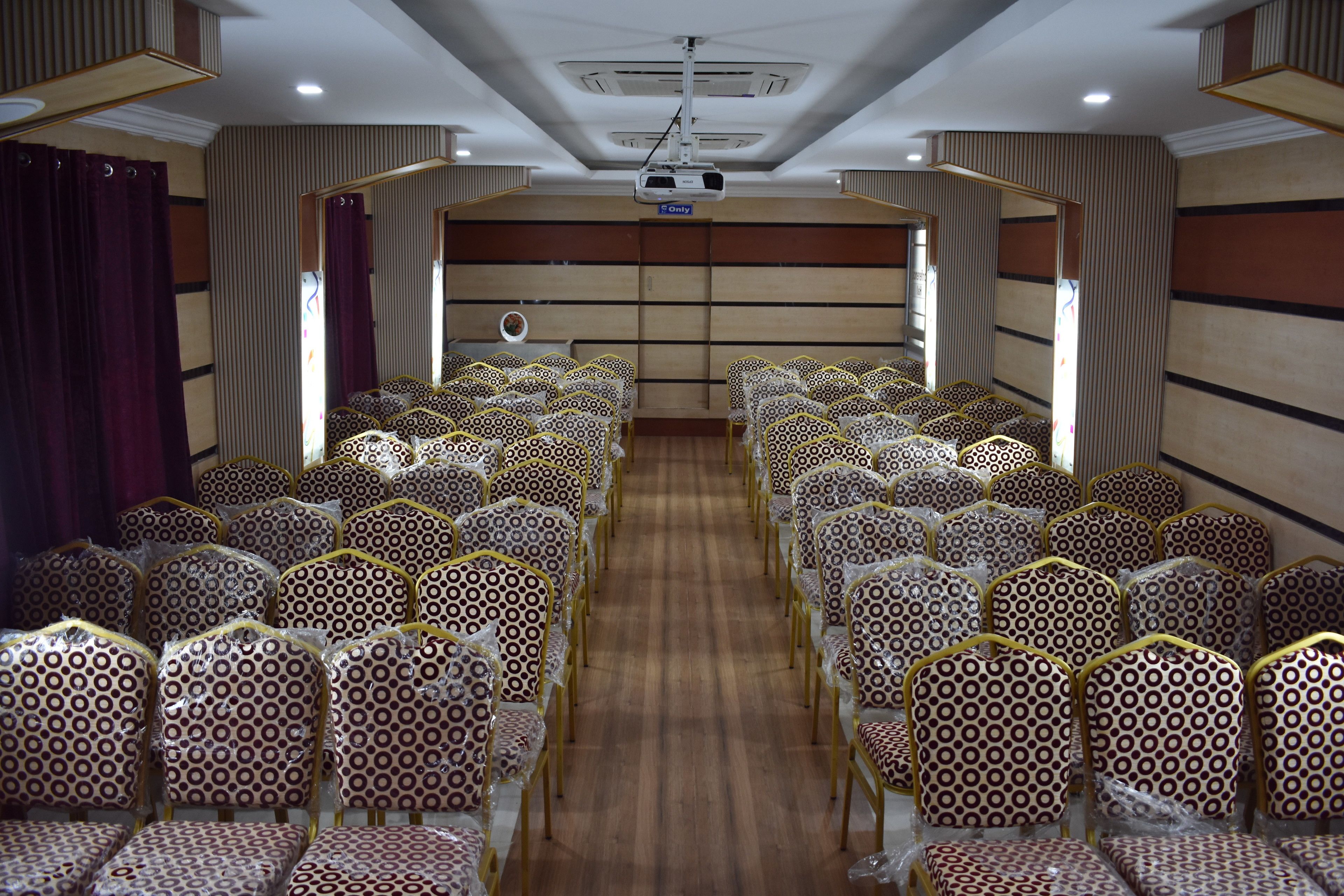 A conference room with rows of patterned chairs, a wooden floor, and a projector screen.