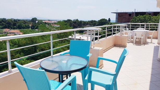 A balcony with a round glass table and blue chairs, overlooking a cityscape with greenery and buildings.