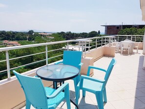 A balcony with a round glass table and blue chairs, overlooking a cityscape with greenery and buildings.