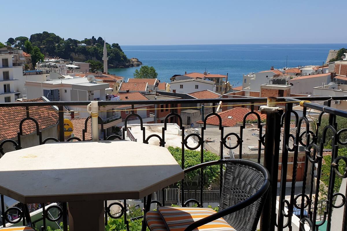 A balcony with a table and chairs, overlooking a coastal town with buildings and a clear view of the sea.